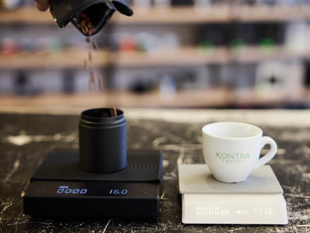 Kaffebønner hældes fra en sort kaffepose og ned i en sort skål der står på en vægt