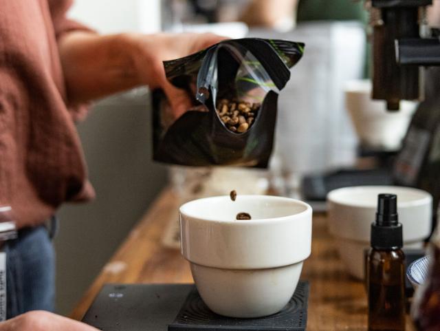 kaffebønner hældes fra en sort kaffepose og ned i en hvid skål der står på en vægt