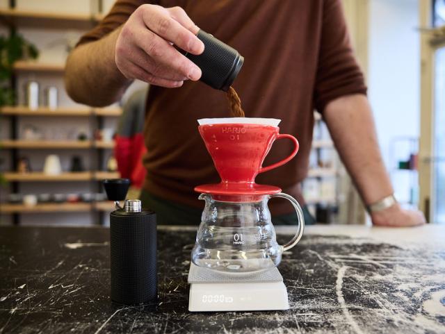 Frisk formalede kaffebønner hældes ned i en rød Hario V60 der står oven på en glasserver og en vægt