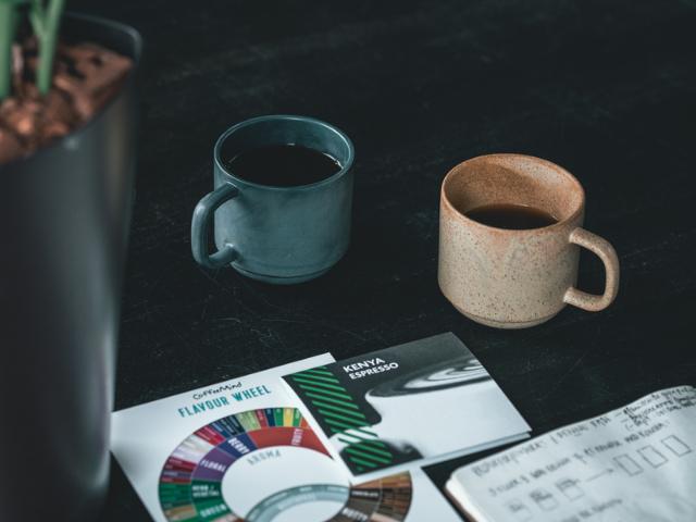 To kaffekopper står på en sort bordplade hvor der også ligger et kaffesmagshjul og andre papirer