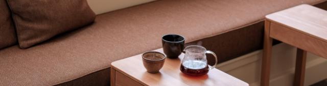 Et lille bord med to kaffekopper og en glaskande med dampende kaffe står foran en lav brun sofa